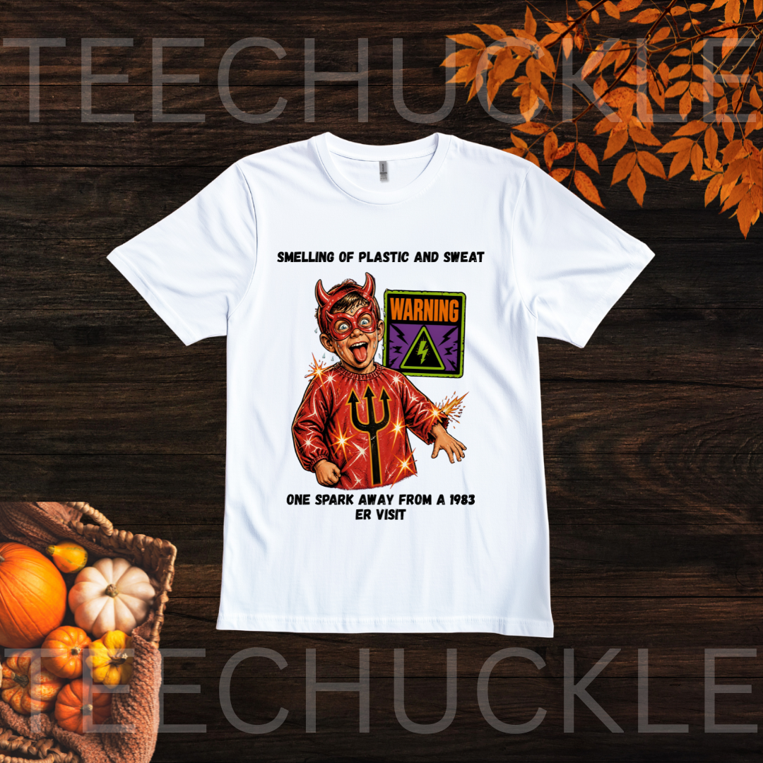 A white short-sleeve graphic t-shirt displayed flat on a dark wood background with autumn-themed props. The graphic features a devil character in a red robe with a black trident, a purple warning sign, and black text reading 'SMELLING OF PLASTIC AND SWEAT' and 'ONE SPARK AWAY FROM A 1985 ER VISIT'.