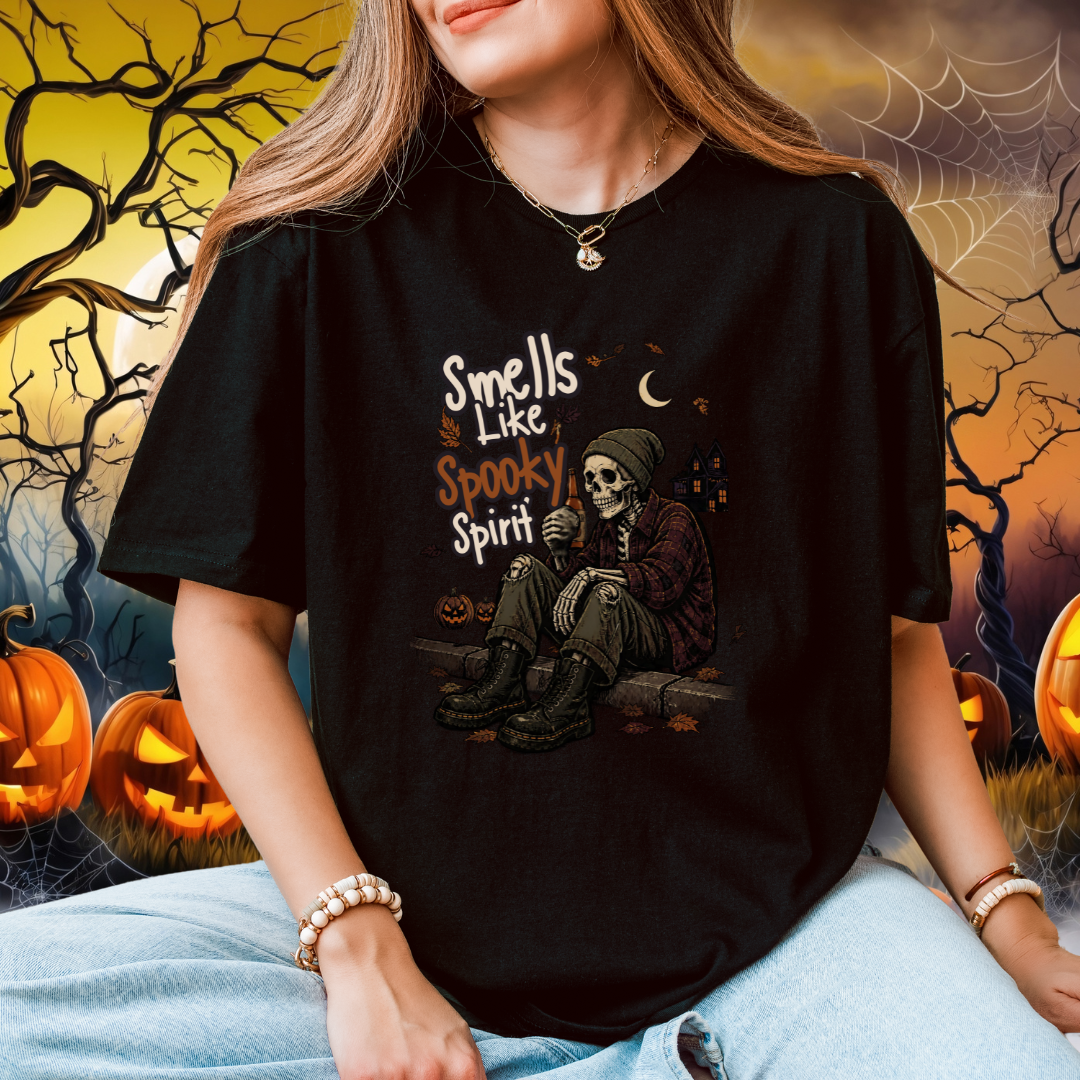 A woman wearing a black graphic t-shirt with a Halloween design that reads 'Smells Like Spooky Spirit' and features a skeleton sitting by a window. The background is a spooky, dark, and eerie Halloween-themed scene with jack-o'-lanterns, bare trees, and cobwebs.