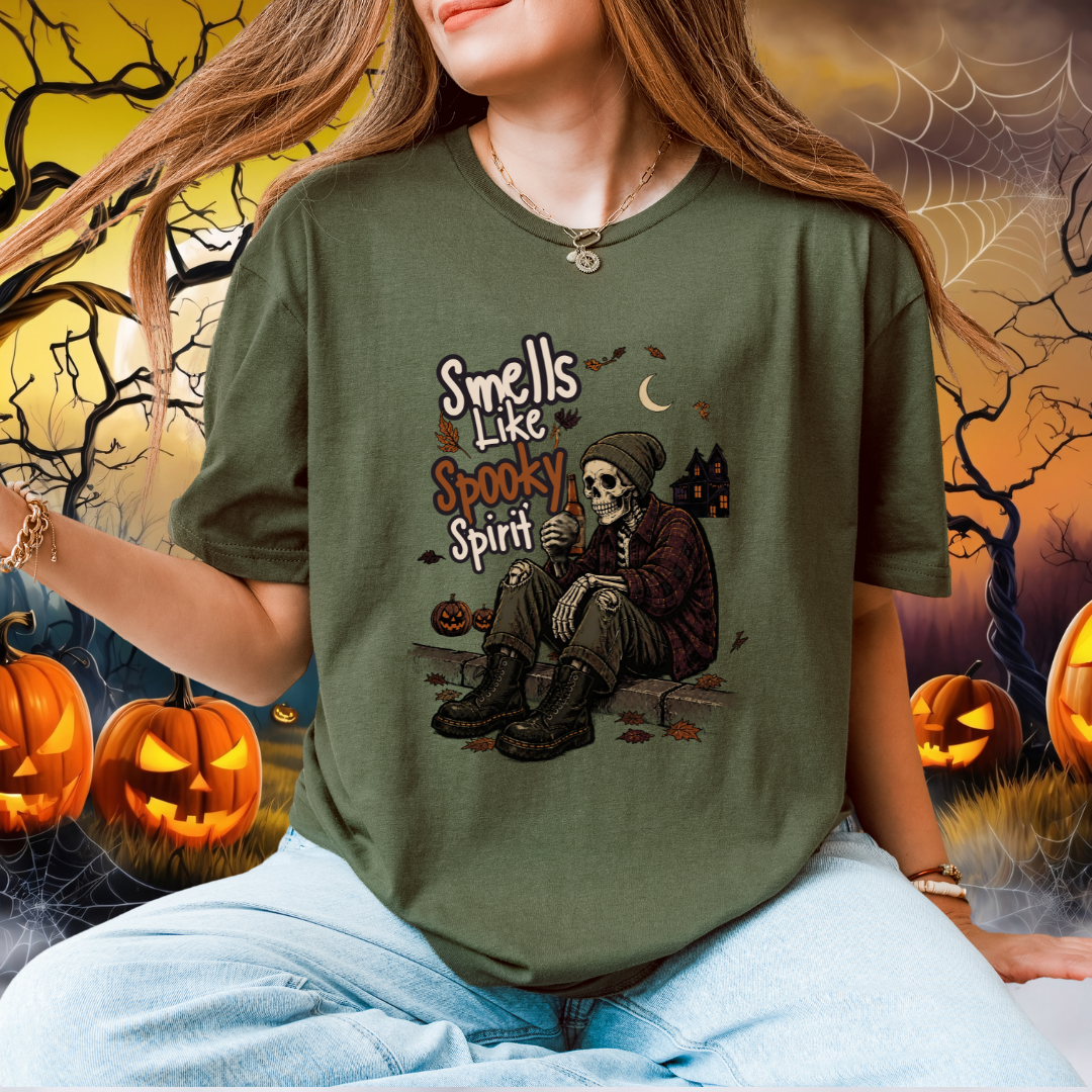 A woman wearing a green graphic t-shirt with a Halloween design of a skeleton sitting on a kerb. The background is a spooky, digitally composited Halloween scene with pumpkins, cobwebs, and a full moon.