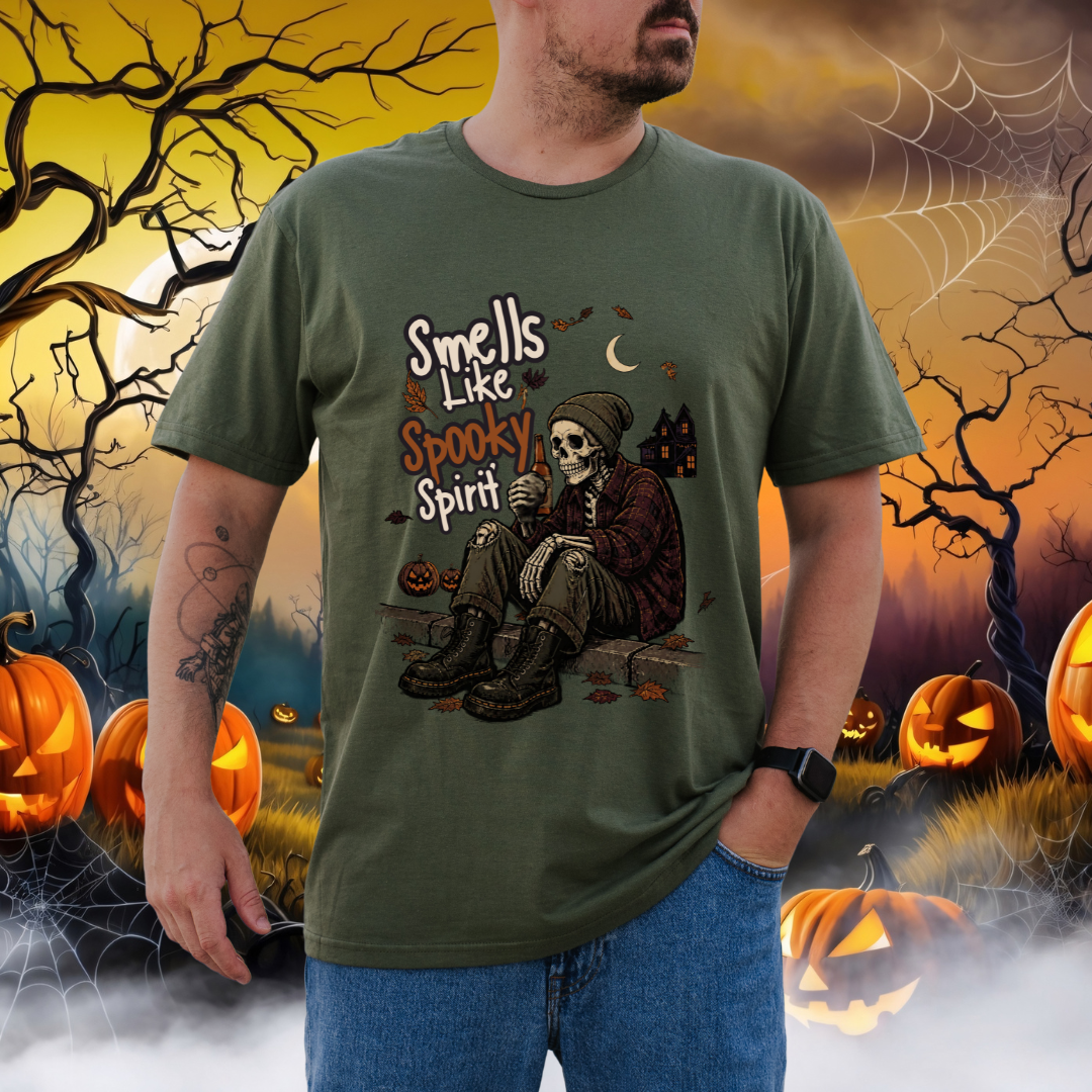 A man wearing a green graphic t-shirt with a Halloween design that reads 'Smells Like Spooky Spirit' next to an illustration of a skeleton sitting in a haunted house. The background is a spooky Halloween scene with pumpkins, cobwebs, and bare trees.