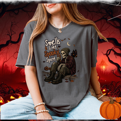 A woman wearing a dark gray graphic t-shirt with a skeleton sitting and the text 'Smells Like Spooky Spirit'. The background is a spooky Halloween scene with pumpkins, bare trees, and a red sky.