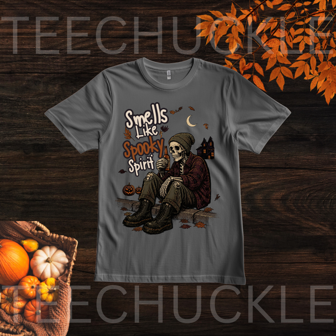 A gray short-sleeve graphic T-shirt laid flat on a dark wood background with autumn props. The shirt features a Halloween-style illustration of a seated skeleton holding a bottle, with the text 'Smells Like Spooky Spirit' and small details including a crescent moon, haunted house, pumpkins, and leaves.