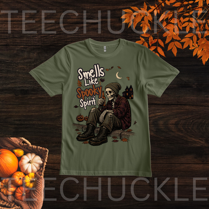 A green graphic t-shirt featuring a skeleton sitting on the ground with the text 'Smells Like Spooky Spirit' in orange and white lettering. The shirt is displayed on a dark wood background with autumn-themed props including pumpkins and orange leaves