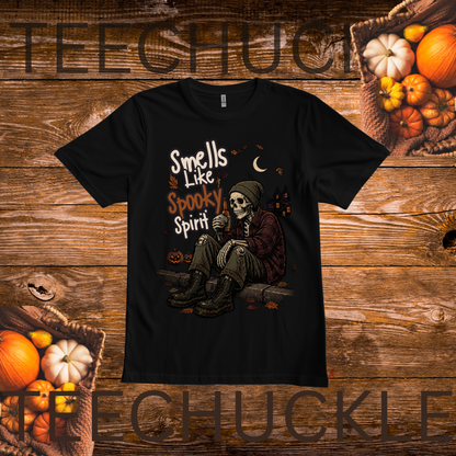 A black short-sleeve graphic T-shirt laid flat on a rustic wooden background with pumpkins and autumn props around it. The shirt features a Halloween-themed illustration of a seated skeleton figure with the text 'Smells Like Spooky Spirit.