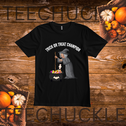 A black short-sleeve graphic T-shirt laid flat on a rustic wooden background with pumpkins and autumn props. The shirt has a printed design of a witch stirring a cauldron of candy, with the words “TRICK OR TREAT CHAMPION” above the illustration.