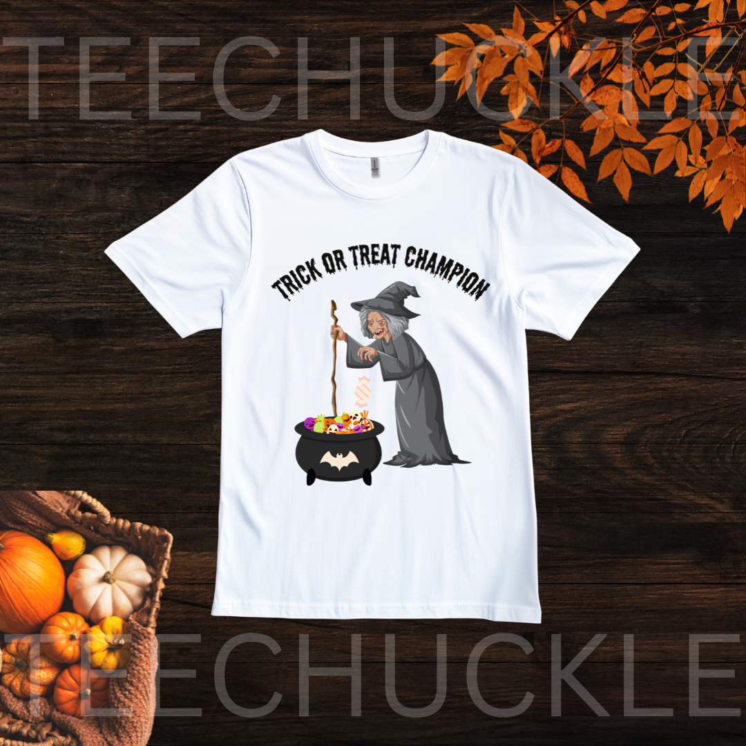 A white short-sleeve graphic T-shirt laid flat on a dark wood background with autumn props. The shirt has black text reading 'TRICK OR TREAT CHAMPION' above a cartoon witch in a gray robe standing by a cauldron filled with colorful treats.