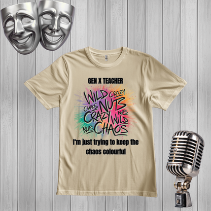 A sand graphic t-shirt displayed on a gray wood plank background, flanked by silver comedy masks on the top left and a vintage-style microphone on the bottom right. The t-shirt features a colorful, hand-drawn style graphic with the words 'WILD CRAZY CHAOS NUTS' in black and multicolor lettering, with 'GEN X TEACHER' above and 'I'm just trying to keep the chaos colourful' below.