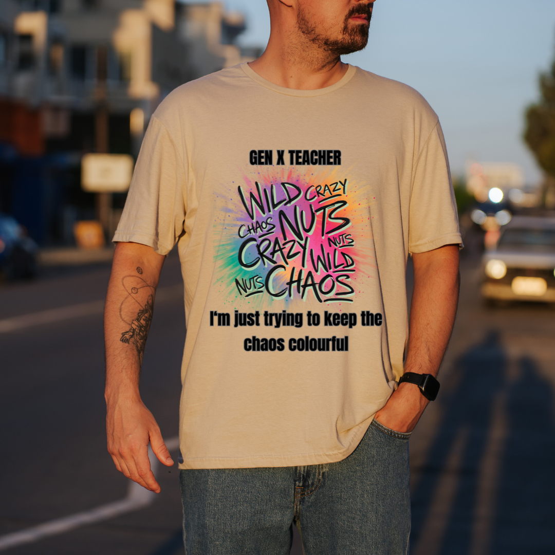 A man wearing a SAND graphic t-shirt with a colorful 'Gen X Teacher' design, standing on a blurred street background at sunset.