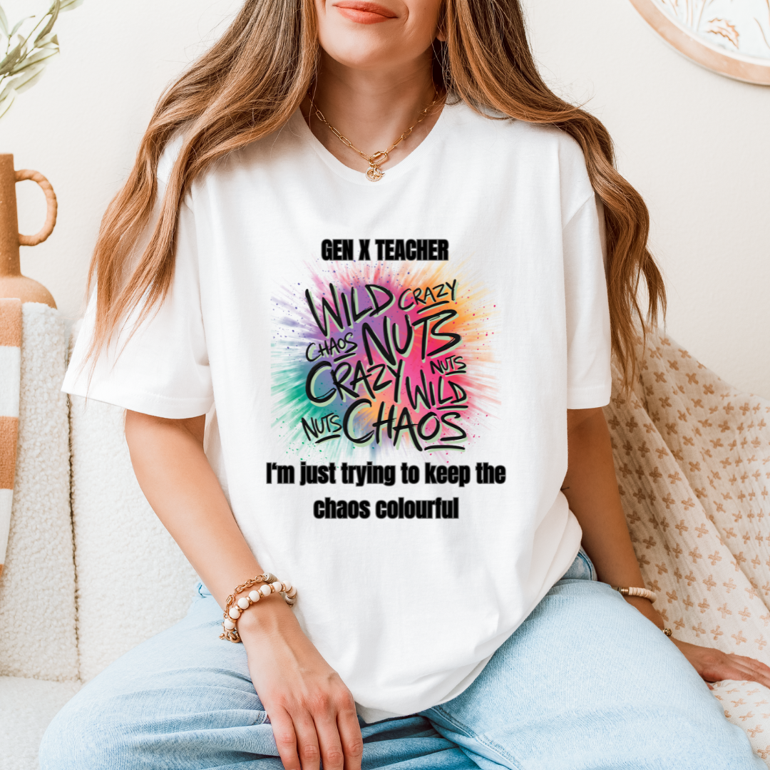 A woman wearing a white graphic T-shirt with a colorful hand-drawn style print that reads 'GEN X TEACHER' and 'I'm just trying to keep the chaos colourful'. The photo is taken in a cozy indoor setting with a textured sofa and neutral decor in the background.
