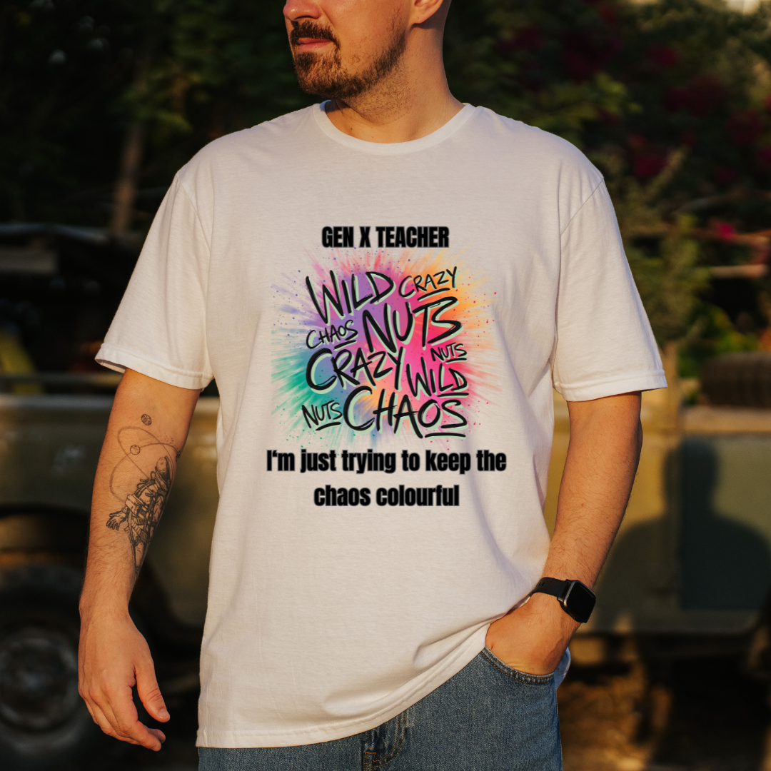 A man wearing a white graphic t-shirt with a colorful 'Gen X Teacher' slogan print, standing outdoors in front of a blurred military vehicle.