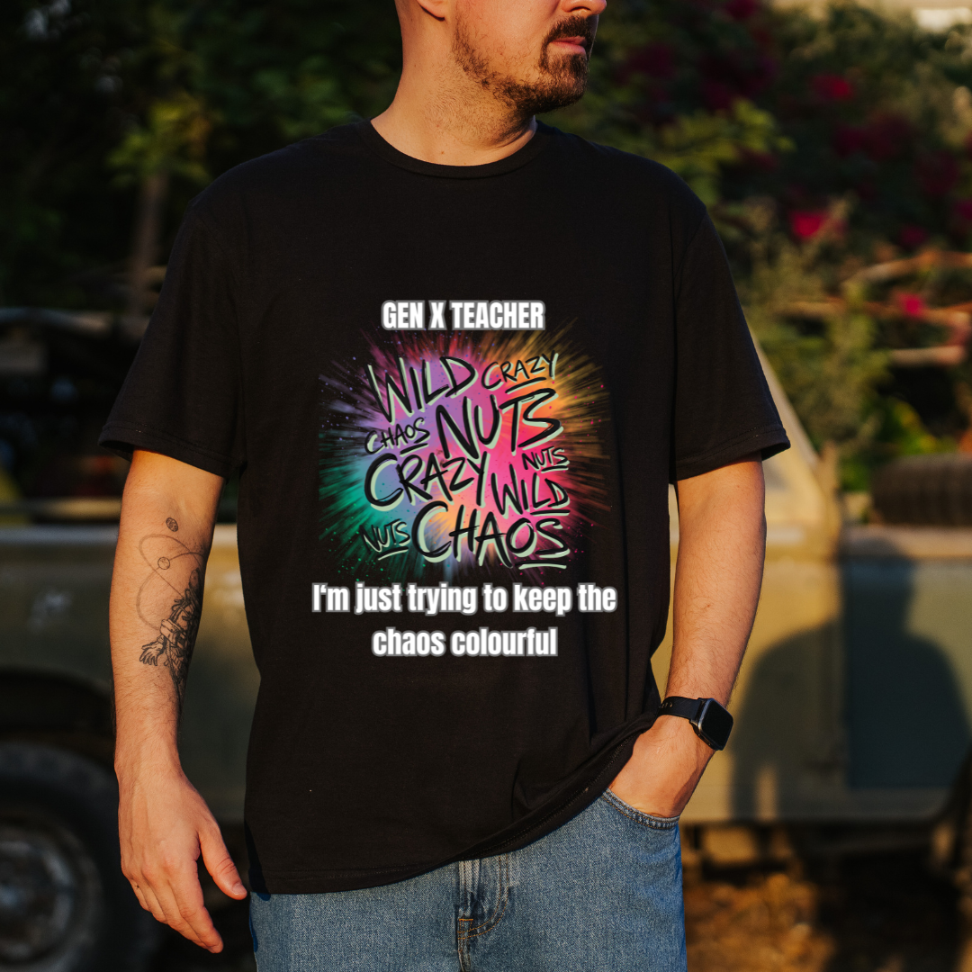A man wearing a black graphic t-shirt with a colorful 'Gen X Teacher im just trying to keeP the chaos colourful ' design, standing outdoors in front of a blurred vehicle and foliage.