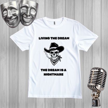 A white short-sleeve graphic T-shirt laid flat on a gray wood plank background, with a black printed skull wearing a cowboy hat and bandana and the text 'LIVING THE DREAM' above and 'THE DREAM IS A NIGHTMARE' below. Silver comedy and tragedy masks appear in the upper left, and a vintage-style microphone appears in the lower right.