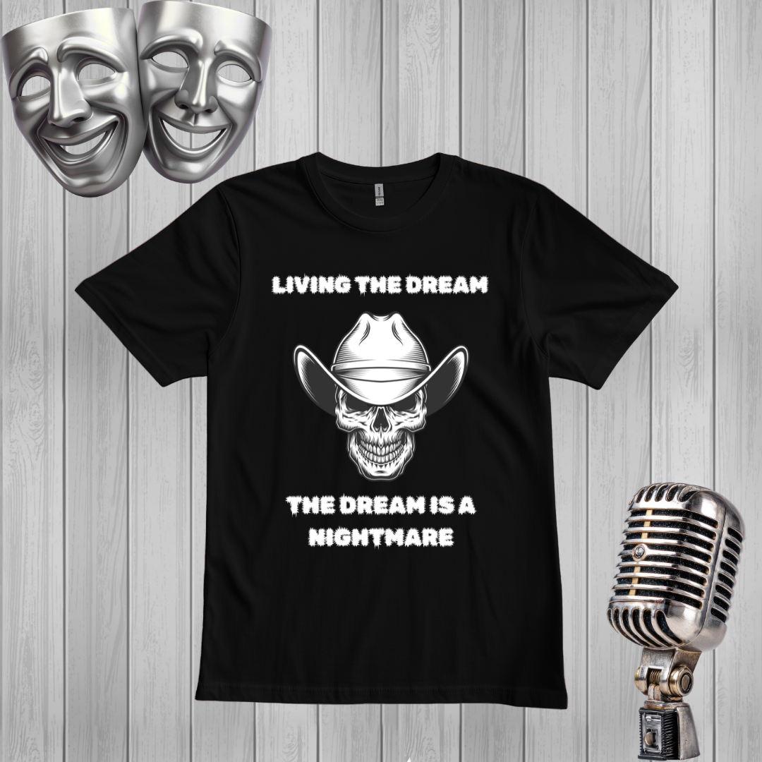 A black short-sleeve graphic T-shirt laid flat on a light gray wood-plank background, with a white skull wearing a cowboy hat and the text 'LIVING THE DREAM THE DREAM IS A NIGHTMARE.' Two silver comedy/tragedy masks appear in the upper left, and a vintage silver microphone appears in the lower right.