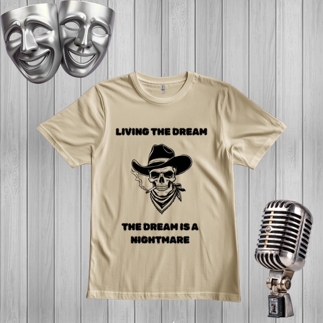 A sand graphic t-shirt displayed on a gray wood-plank background, flanked by silver theater masks and a vintage-style microphone. The t-shirt features a black graphic of a skull wearing a cowboy hat and smoking a cigar, with the text 'LIVING THE DREAM THE DREAM IS A NIGHTMARE'.
