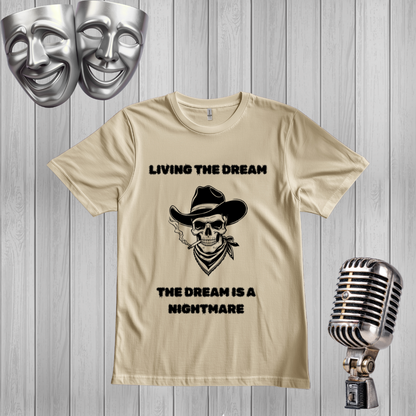 A sand graphic t-shirt displayed on a gray wood-plank background, flanked by silver theater masks and a vintage-style microphone. The t-shirt features a black graphic of a skull wearing a cowboy hat and smoking a cigar, with the text 'LIVING THE DREAM THE DREAM IS A NIGHTMARE'.