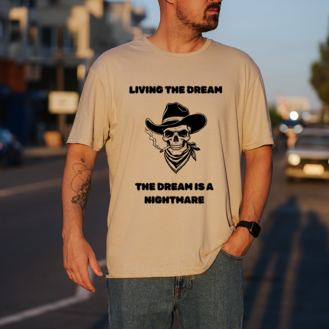 A man wearing a sand graphic t-shirt with a black print of a skull in a cowboy hat and the text 'LIVING THE DREAM THE DREAM IS A NIGHTMARE', standing on a blurred street at sunset.