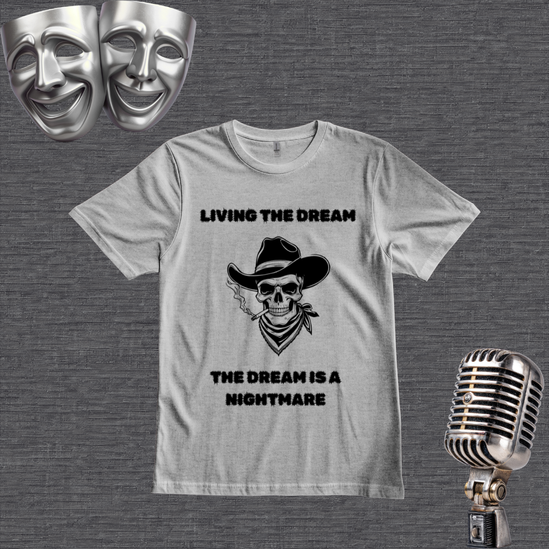 A gray graphic t-shirt featuring a black print of a cowboy skull wearing a hat and bandana, with the text 'LIVING THE DREAM' above and 'THE DREAM IS A NIGHTMARE' below. The shirt is displayed on a textured gray background with vintage silver theater props (a comedy and tragedy mask and a microphone) in the corners.