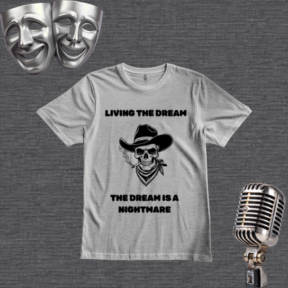 A gray graphic t-shirt featuring a black print of a cowboy skull wearing a hat and bandana, with the text 'LIVING THE DREAM' above and 'THE DREAM IS A NIGHTMARE' below. The shirt is displayed on a textured gray background with vintage silver theater props (a comedy and tragedy mask and a microphone) in the corners.