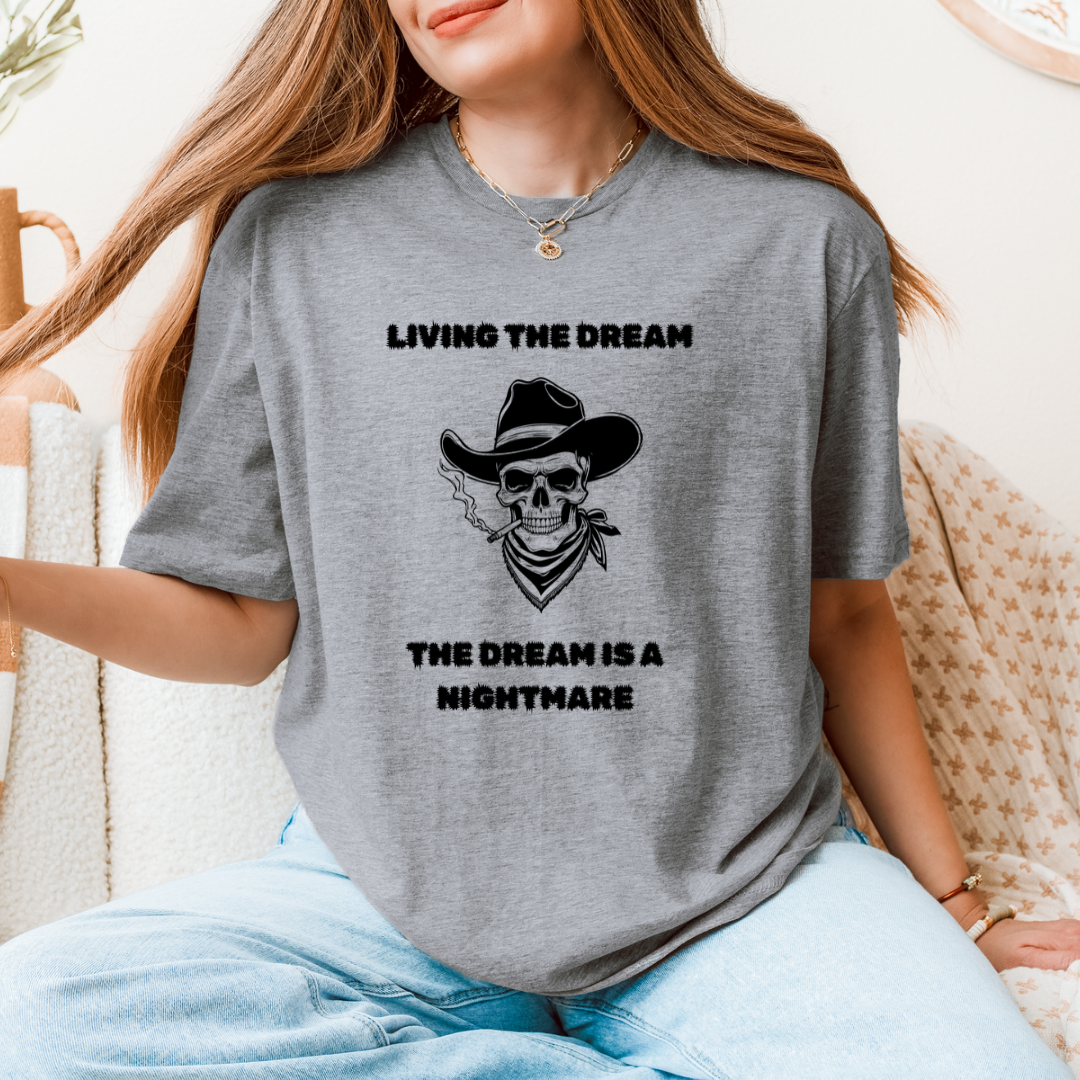 A woman wearing a gray graphic t-shirt with a black printed design of a skull in a cowboy hat smoking a cigar, with the text 'LIVING THE DREAM THE DREAM IS A NIGHTMARE'.
