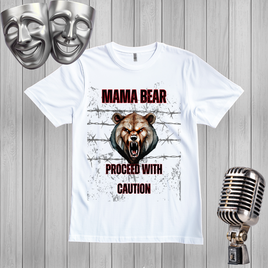 A white graphic t-shirt featuring a roaring bear design with the text 'MAMA BEAR PROCEED WITH CAUTION' and barbed wire, displayed on a gray wood plank background with silver theater masks and a vintage microphone prop.