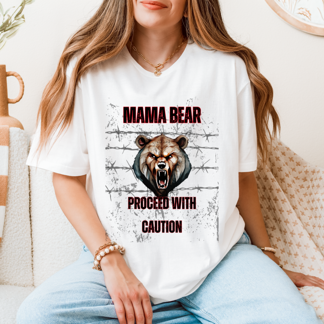 A woman wearing a white graphic t-shirt with a 'MAMA BEAR PROCEED WITH CAUTION' design featuring a roaring bear and barbed wire, seated in a cozy indoor setting.