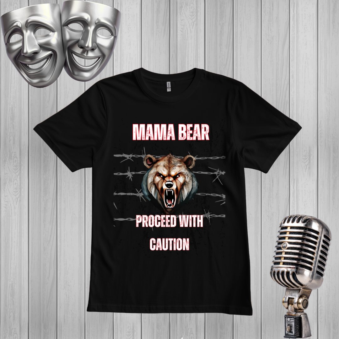 A black graphic t-shirt featuring a roaring bear design with the text 'MAMA BEAR' and 'PROCEED WITH CAUTION' in pink. The shirt is displayed on a gray wood-plank background with silver comedy masks and a vintage microphone as props.
