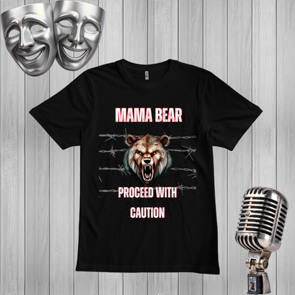 A black graphic t-shirt featuring a roaring bear design with the text 'MAMA BEAR' and 'PROCEED WITH CAUTION' in pink. The shirt is displayed on a gray wood-plank background with silver comedy masks and a vintage microphone as props.