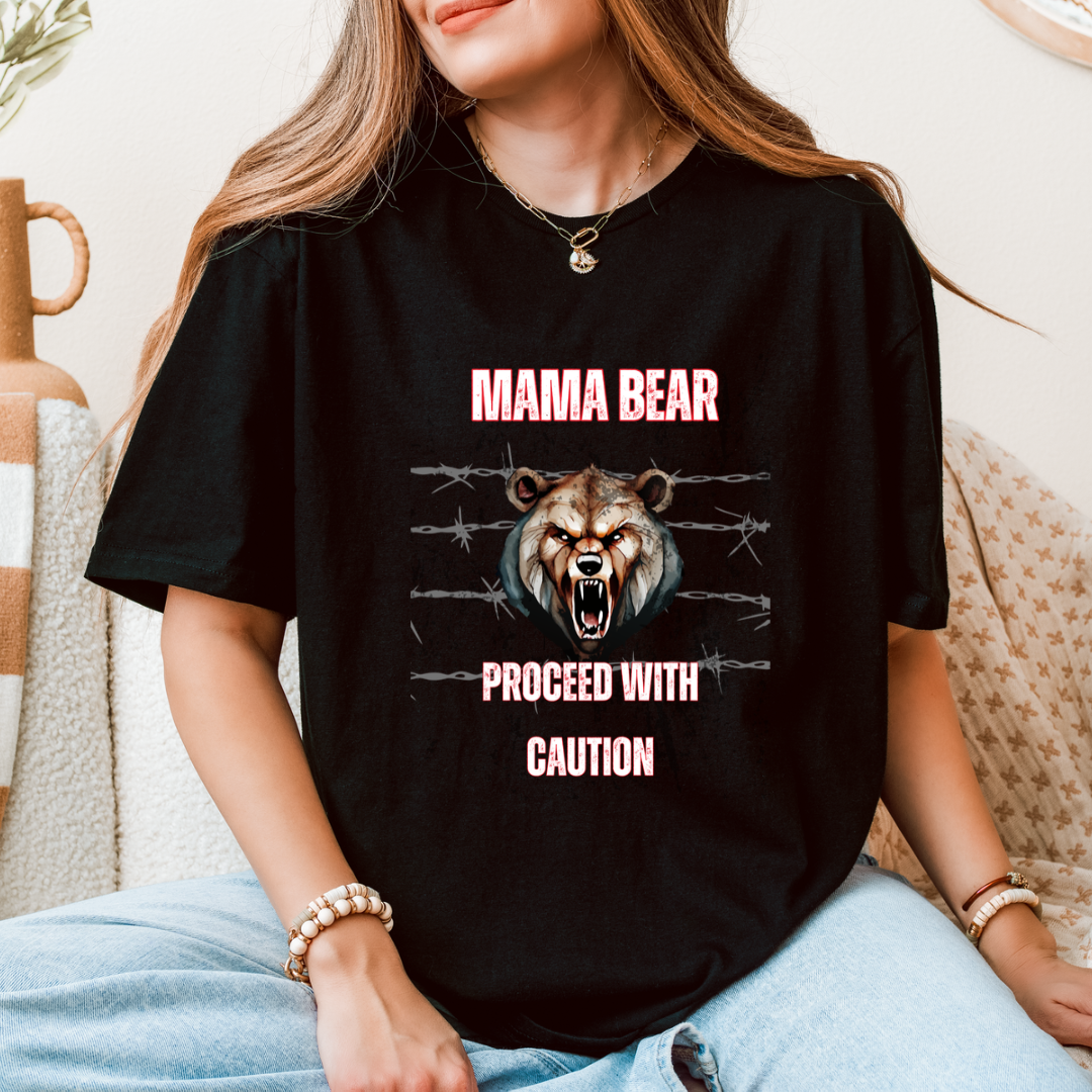 A woman wearing a black graphic t-shirt with a roaring bear design and the text 'MAMA BEAR PROCEED WITH CAUTION' in pink, sitting on a beige couch.