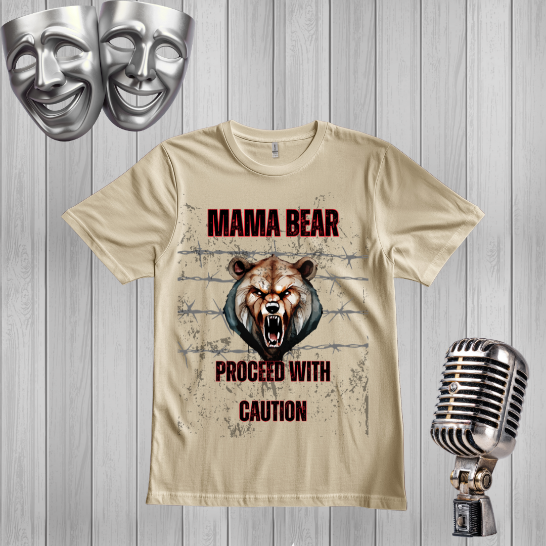 A sand graphic t-shirt featuring a roaring bear design with the text 'MAMA BEAR PROCEED WITH CAUTION'. The t-shirt is displayed on a gray wood-plank background, styled with a silver microphone on the right and two silver comedy and tragedy masks on the top left.
