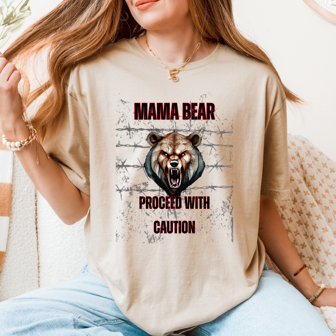 A woman wearing a beige graphic t-shirt with a roaring bear design and the text 'MAMA BEAR PROCEED WITH CAUTION' in dark red letters, paired with light blue jeans in a cozy indoor setting.