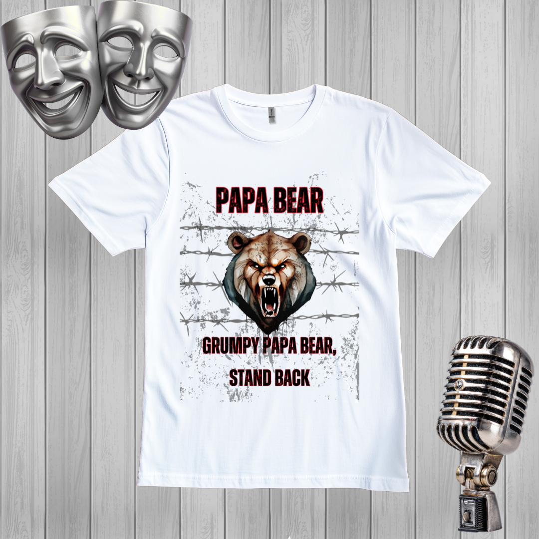 A white graphic t-shirt featuring a grumpy bear design with the text 'PAPA BEAR' and 'GRUMPY PAPA BEAR, STAND BACK'. The shirt is displayed on a gray wood panel background with silver comedy masks in the top-left corner and a vintage silver microphone on the bottom-right.