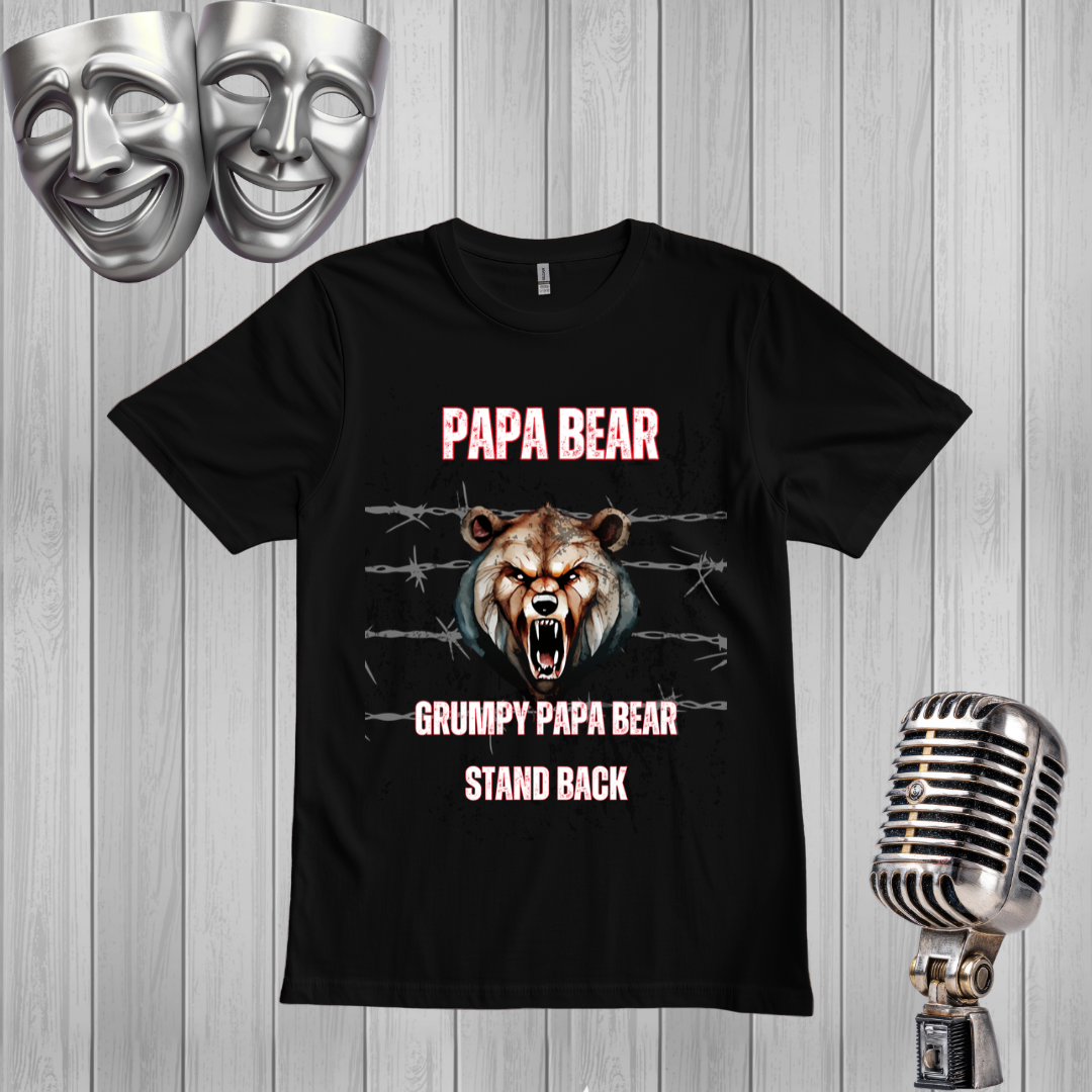 A black graphic t-shirt featuring a grumpy bear design with the text 'PAPA BEAR' and 'GRUMPY PAPA BEAR STAND BACK'. The shirt is displayed on a light gray wood-plank background, flanked by silver props including two comedy masks at the top left and a vintage-style microphone at the bottom right.
