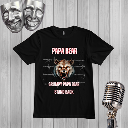 A black graphic t-shirt featuring a grumpy bear design with the text 'PAPA BEAR' and 'GRUMPY PAPA BEAR STAND BACK'. The shirt is displayed on a light gray wood-plank background, flanked by silver props including two comedy masks at the top left and a vintage-style microphone at the bottom right.