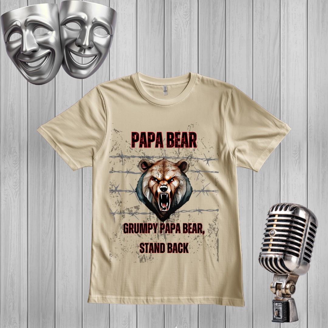 A sand graphic t-shirt featuring a roaring bear design with the text 'PAPA BEAR' and 'GRUMPY PAPA BEAR, STAND BACK'. The shirt is displayed on a gray wood-plank background alongside decorative props, including two silver comedy masks at the top left and a vintage silver microphone at the bottom right.