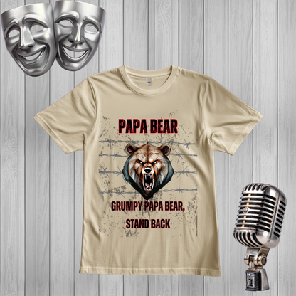 A sand graphic t-shirt featuring a roaring bear design with the text 'PAPA BEAR' and 'GRUMPY PAPA BEAR, STAND BACK'. The shirt is displayed on a gray wood-plank background alongside decorative props, including two silver comedy masks at the top left and a vintage silver microphone at the bottom right.