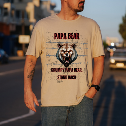 A man wearing a sand graphic t-shirt with a grumpy bear design and the text 'PAPA BEAR GRUMPY PAPA BEAR STAND BACK', standing on a blurred street at sunset.