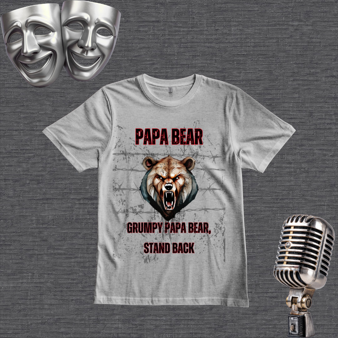 A gray graphic t-shirt featuring a grumpy bear design with the text 'PAPA BEAR' and 'GRUMPY PAPA BEAR, STAND BACK'. The t-shirt is displayed on a dark gray textured background alongside thematic props, including two silver comedy masks in the top-left and a vintage silver microphone in the bottom-right.