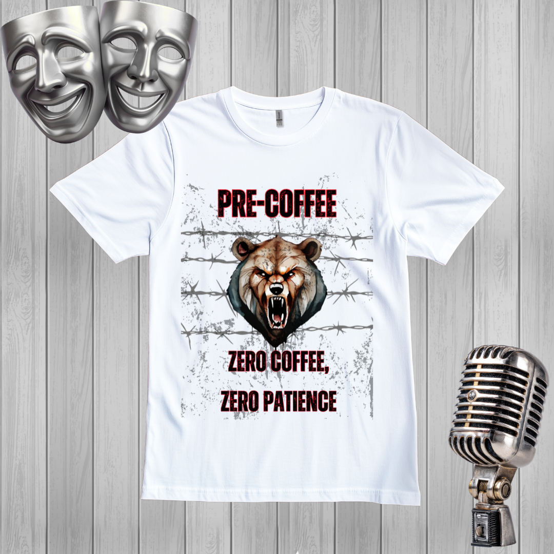 A white graphic t-shirt displayed on a gray wood plank background, flanked by silver comedy and tragedy masks on the top left and a vintage silver microphone on the bottom right. The t-shirt features a graphic of an angry brown bear head with the text 'PRE-COFFEE ZERO COFFEE, ZERO PATIENCE' in dark red and white.