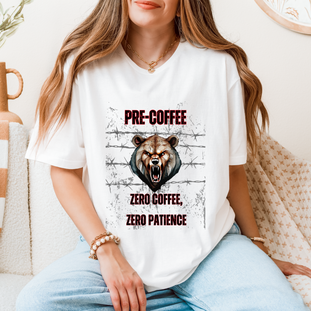 A woman wearing a white graphic t-shirt with a roaring bear and the text 'PRE-COFFEE ZERO COFFEE, ZERO PATIENCE' in a cozy indoor setting.