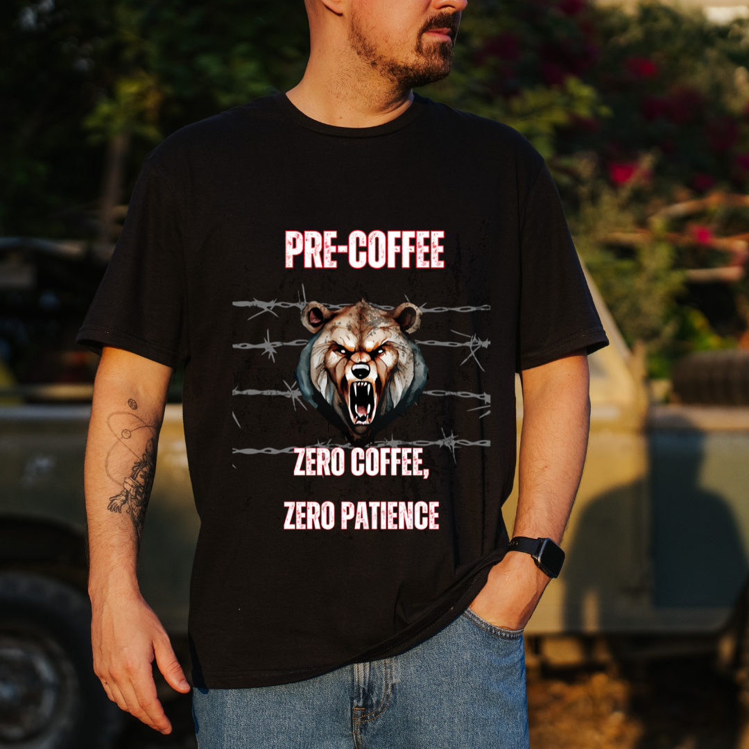 A man wearing a black graphic t-shirt with a roaring bear design and the text 'PRE-COFFEE ZERO COFFEE, ZERO PATIENCE' in pink, standing outdoors in front of a blurred vehicle and foliage.
