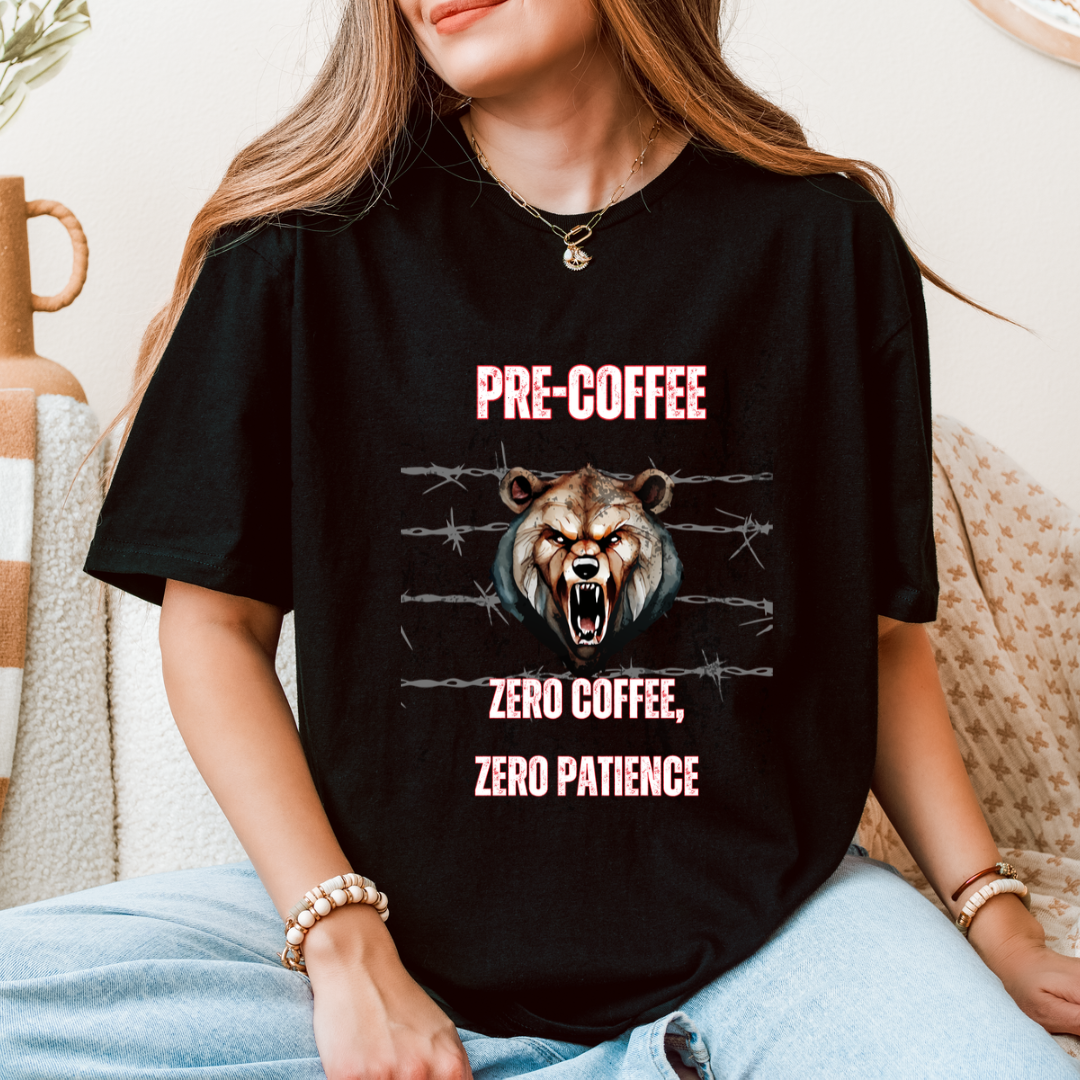 A woman wearing a black graphic t-shirt with a roaring bear design and the text 'PRE-COFFEE ZERO COFFEE, ZERO PATIENCE' in pink, sitting on a beige couch.