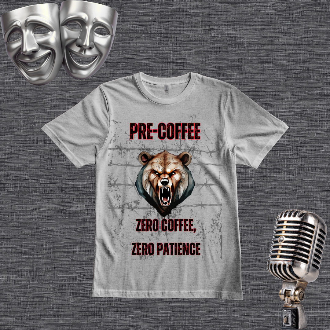 A gray graphic t-shirt featuring a roaring bear design with the text 'PRE COFFEE ZERO COFFEE, ZERO PATIENCE' in red and black. The t-shirt is displayed on a textured gray background alongside thematic props, including two silver comedy masks in the top-left and a vintage silver microphone in the bottom-right.