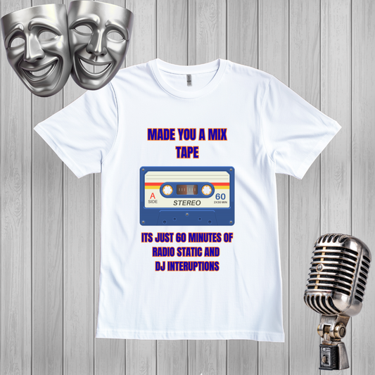 A white graphic t-shirt displayed on a gray wood plank background, flanked by silver comedy masks on the left and a vintage silver microphone on the right. The t-shirt features a retro cassette tape graphic with purple text reading 'MADE YOU A MIX TAPE' and 'ITS JUST 60 MINUTES OF RADIO STATIC AND DJ INTERUPTIONS'.