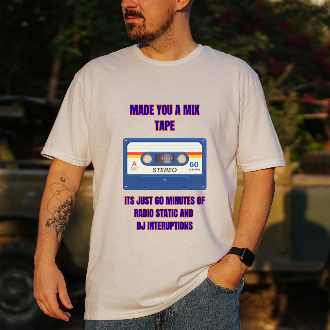 A man wearing a white graphic t-shirt with a retro cassette tape design that reads 'MADE YOU A MIX TAPE' and 'ITS JUST 60 MINUTES OF RADIO STATIC AND DJ INTERRUPTIONS'. The background is a blurred outdoor setting with green foliage and a vehicle.