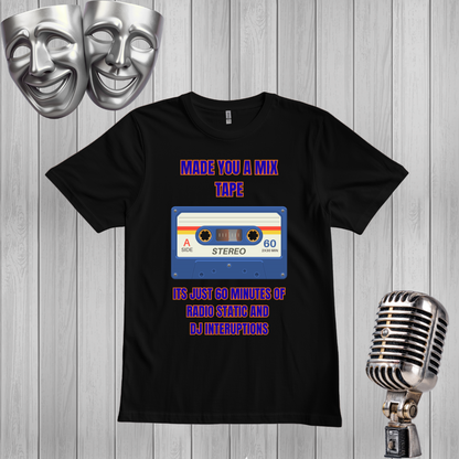 A black graphic t-shirt displayed on a gray wood plank background, flanked by silver comedy masks on the left and a vintage silver microphone on the right. The t-shirt features a retro cassette tape graphic with purple text reading 'MADE YOU A MIX TAPE' and 'ITS JUST 60 MINUTES OF RADIO STATIC AND DJ INTERUPTIONS'.