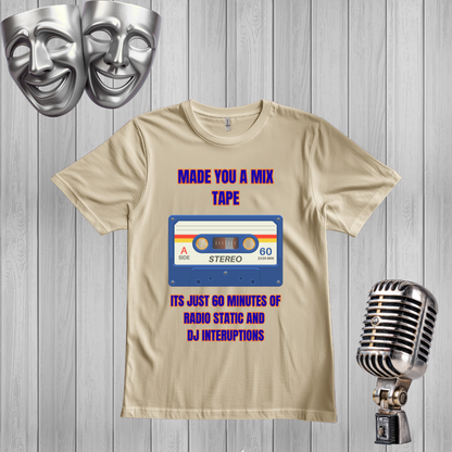 A sand graphic t-shirt displayed on a gray wood plank background, flanked by silver comedy masks on the top left and a vintage silver microphone on the bottom right. The t-shirt features a retro cassette tape graphic with the text 'MADE YOU A MIX TAPE' and 'ITS JUST 60 MINUTES OF RADIO STATIC AND DJ INTERRUPTIONS' in purple and blue.