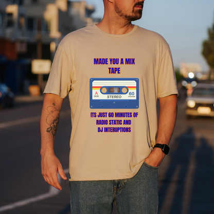 A man wearing a sand graphic t-shirt with a retro cassette tape design and purple text that reads 'MADE YOU A MIX TAPE ITS JUST 60 MINUTES OF RADIO STATIC AND DJ INTERRUPTIONS'. The background is a blurred street scene at sunset.