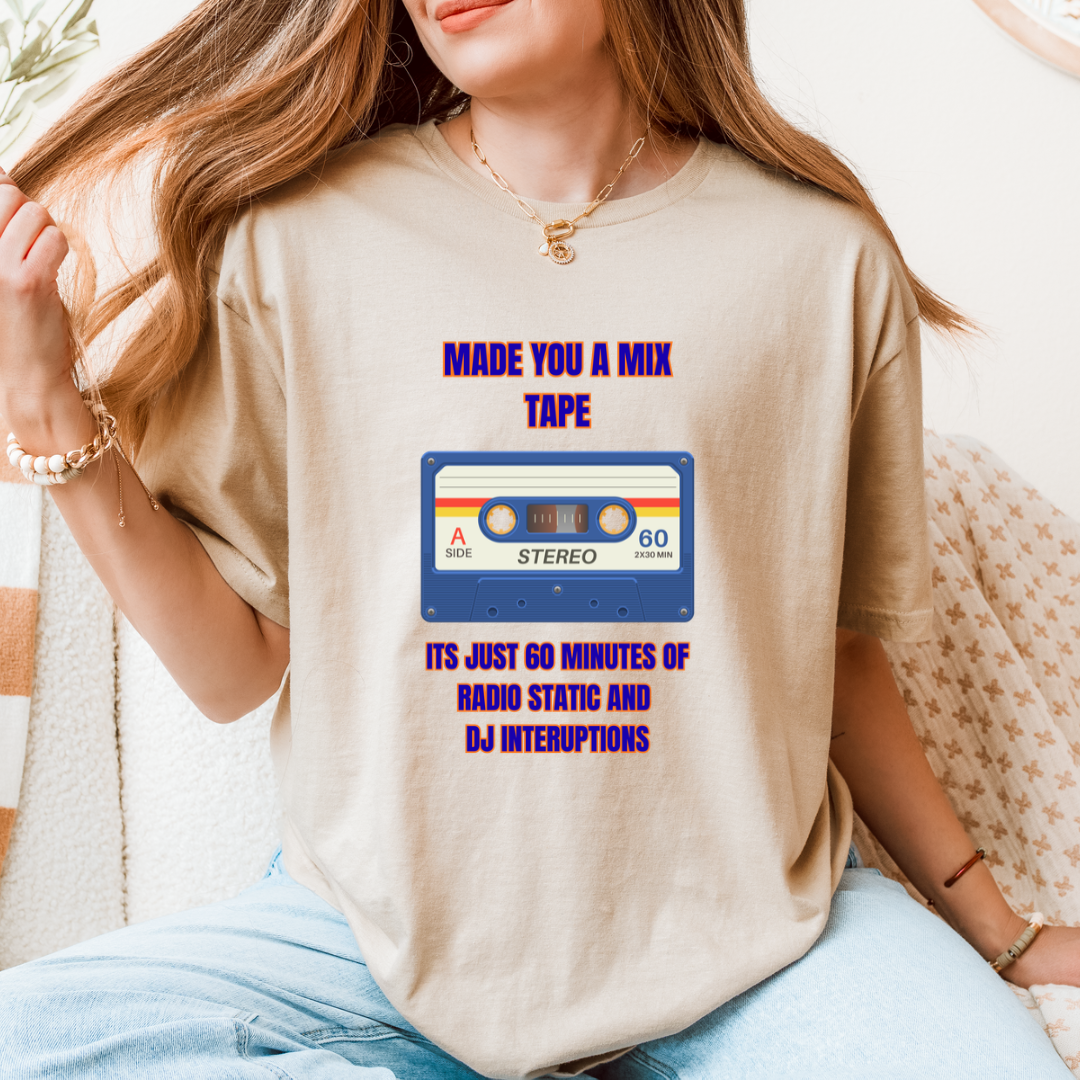 A woman wearing a sand graphic t-shirt with a retro cassette tape design and purple text that reads 'MADE YOU A MIX TAPE ITS JUST 60 MINUTES OF RADIO STATIC AND DJ INTERRUPTIONS'.