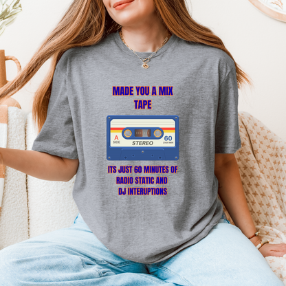 A woman wearing a gray graphic t-shirt with a retro cassette tape design and purple text that reads 'MADE YOU A MIX TAPE ITS JUST 60 MINUTES OF RADIO STATIC AND DJ INTERUPTIONS'. She is sitting on a beige couch in a cozy indoor setting.