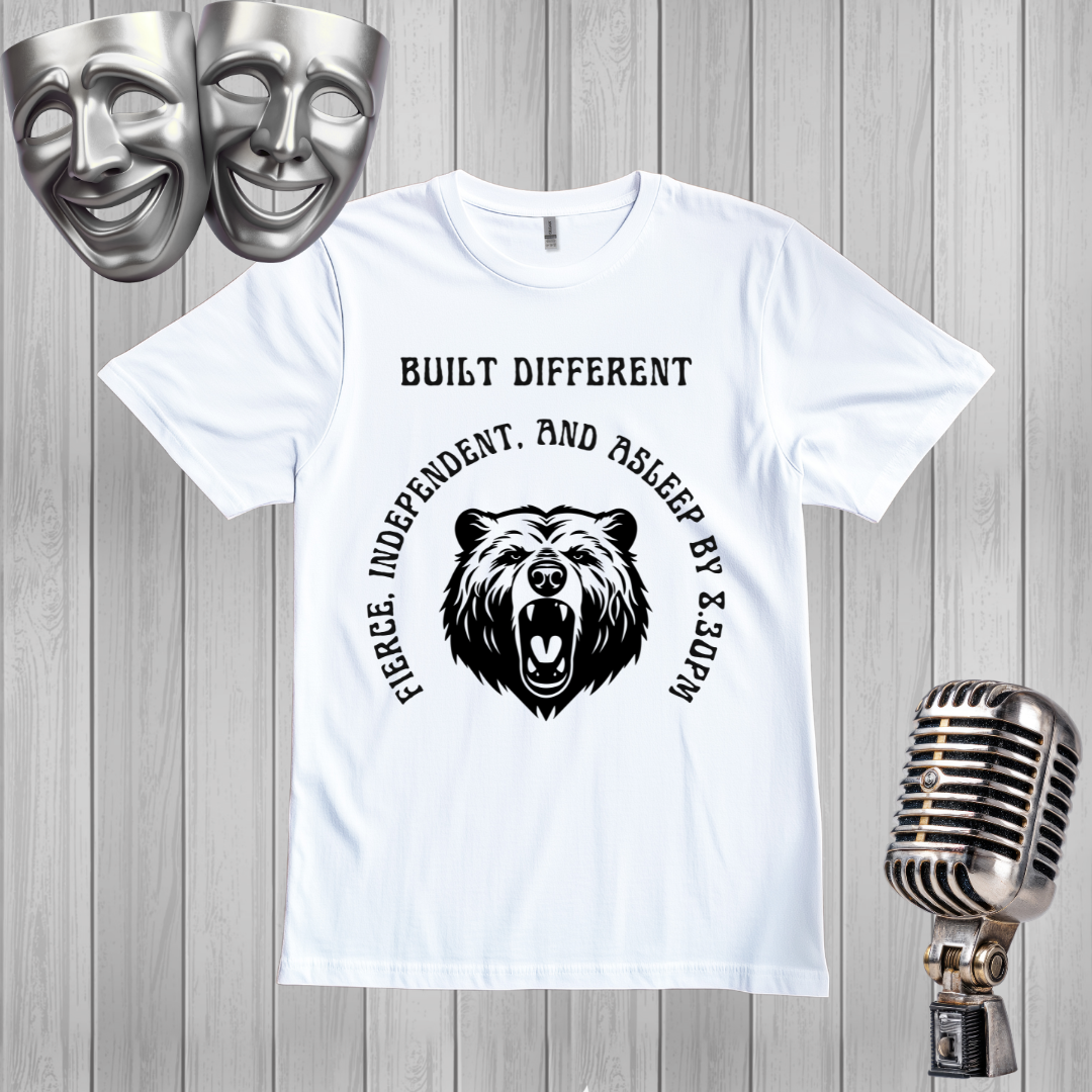 A white short-sleeve graphic T-shirt laid flat on a light gray wood plank background, with black text reading 'BUILT DIFFERENT' above a bear head graphic and curved text around it. Two silver theater masks appear in the top left and a vintage silver microphone appears in the bottom right.